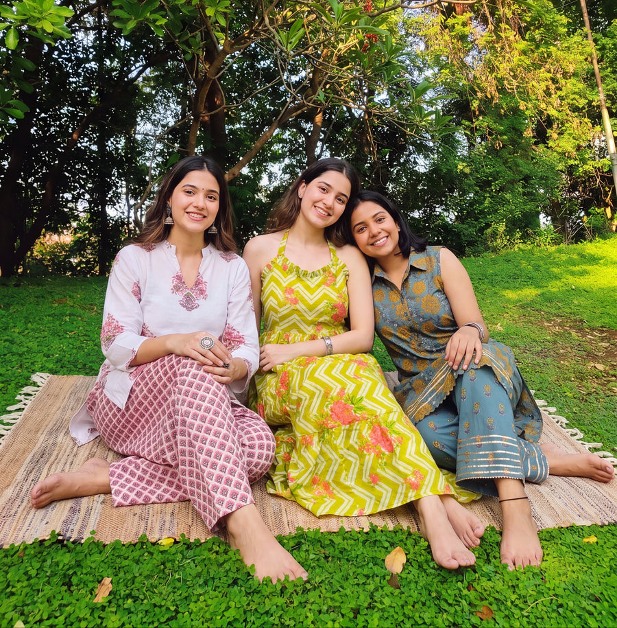 Three women sitting together outdoors on a woven mat on green grass under a tree with red flowers. They are wearing traditional Indian outfits in pink, yellow, and blue-green colors.
